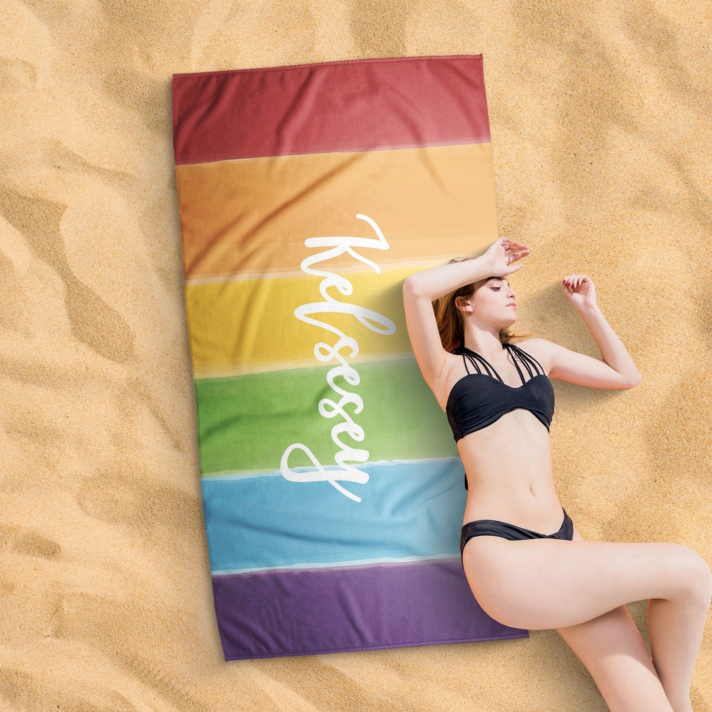 A lady lying on a watercolor rainbow pride design beach towel on a sandy surface