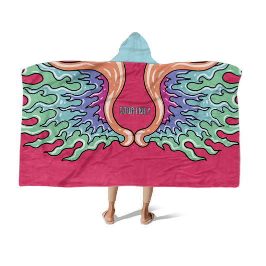 A person wearing a hooded blanket with vibrant, colorful wing designs and the name "Courtney" in the center on a pink background.