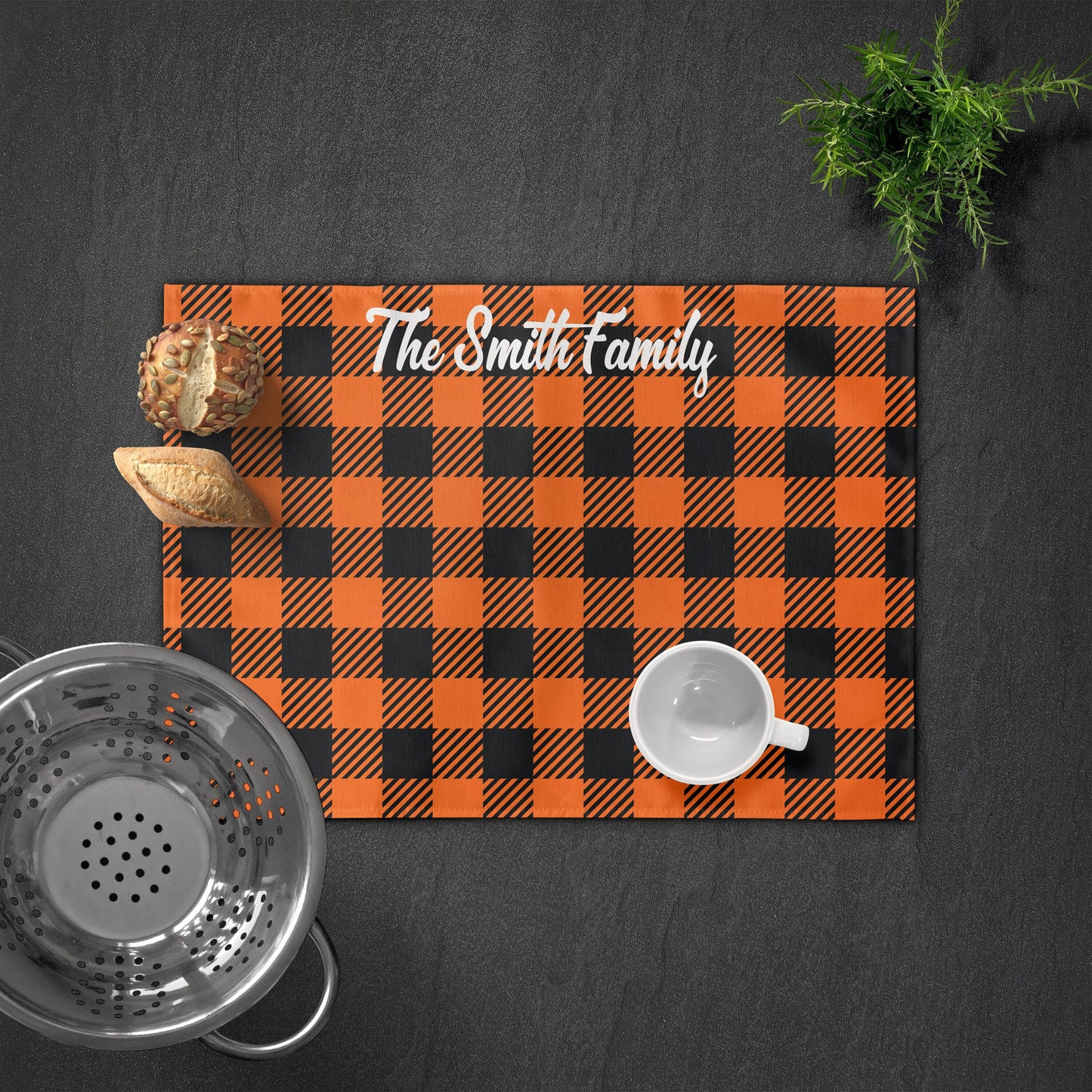 Personalized orange and black placemat with "The Smith Family" text, and bread, colander, cup, and a small plant on a dark surface.