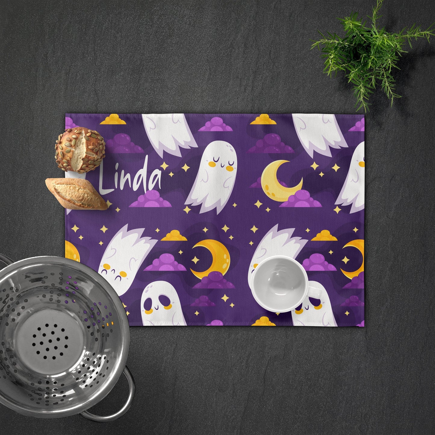 Personalized purple placemat with "Linda," featuring cute white ghosts, moons, and stars. Bread, cup, and colander are nearby