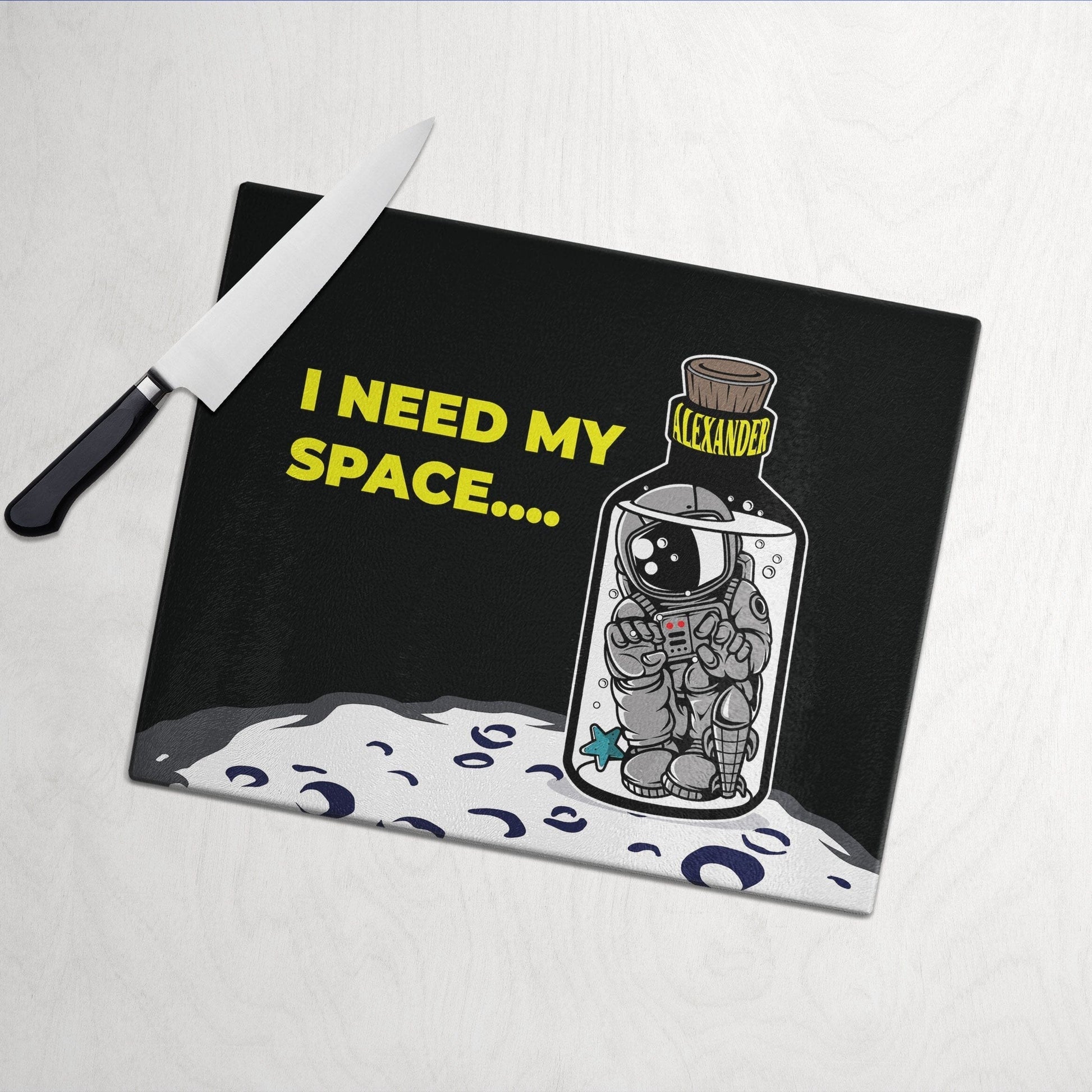 Glass cutting board with "I Need My Space" text, featuring an astronaut in a bottle labeled "Alexander," on a moon surface.