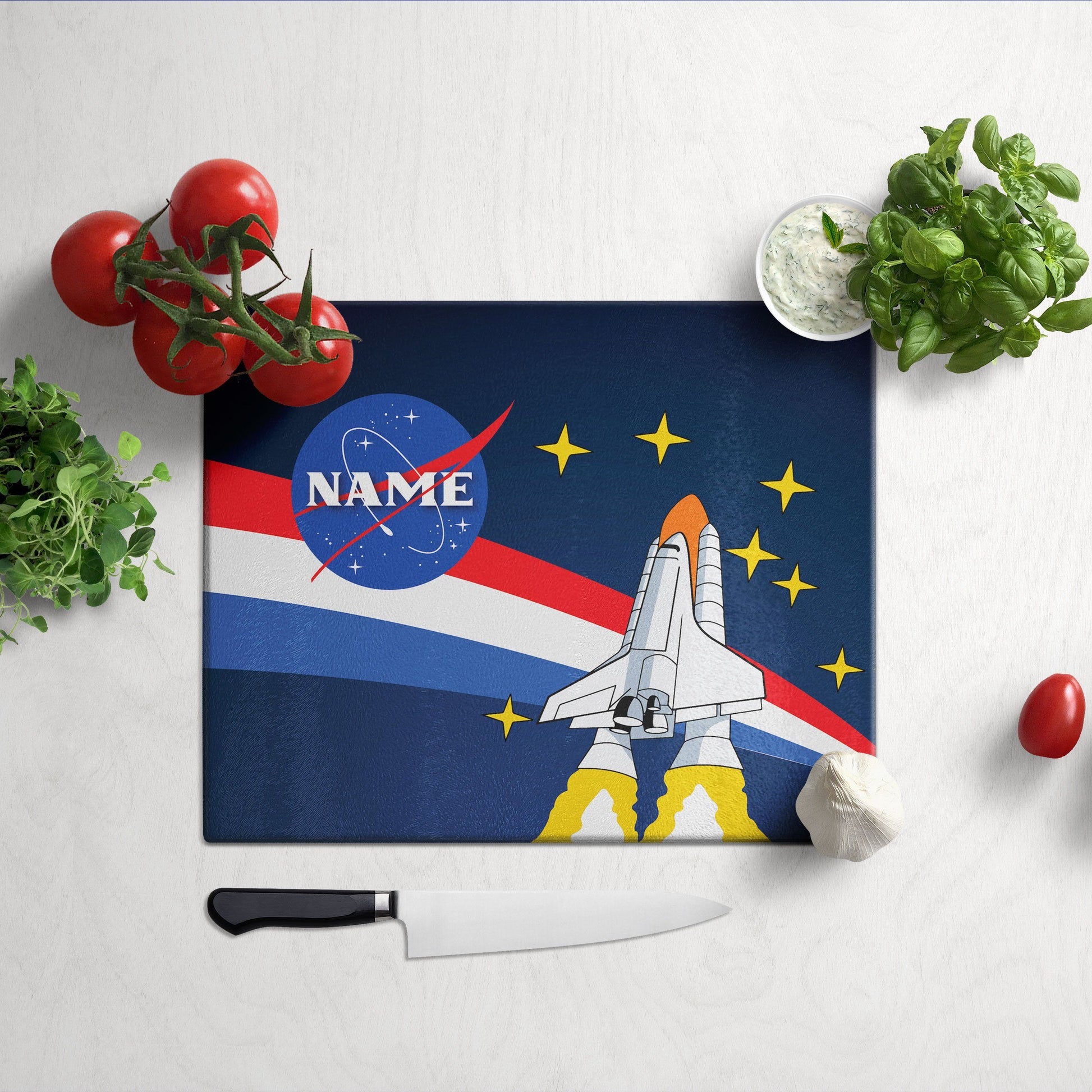 A glass cutting board with nasa launch design on a grey surface and knife & veggies on the side