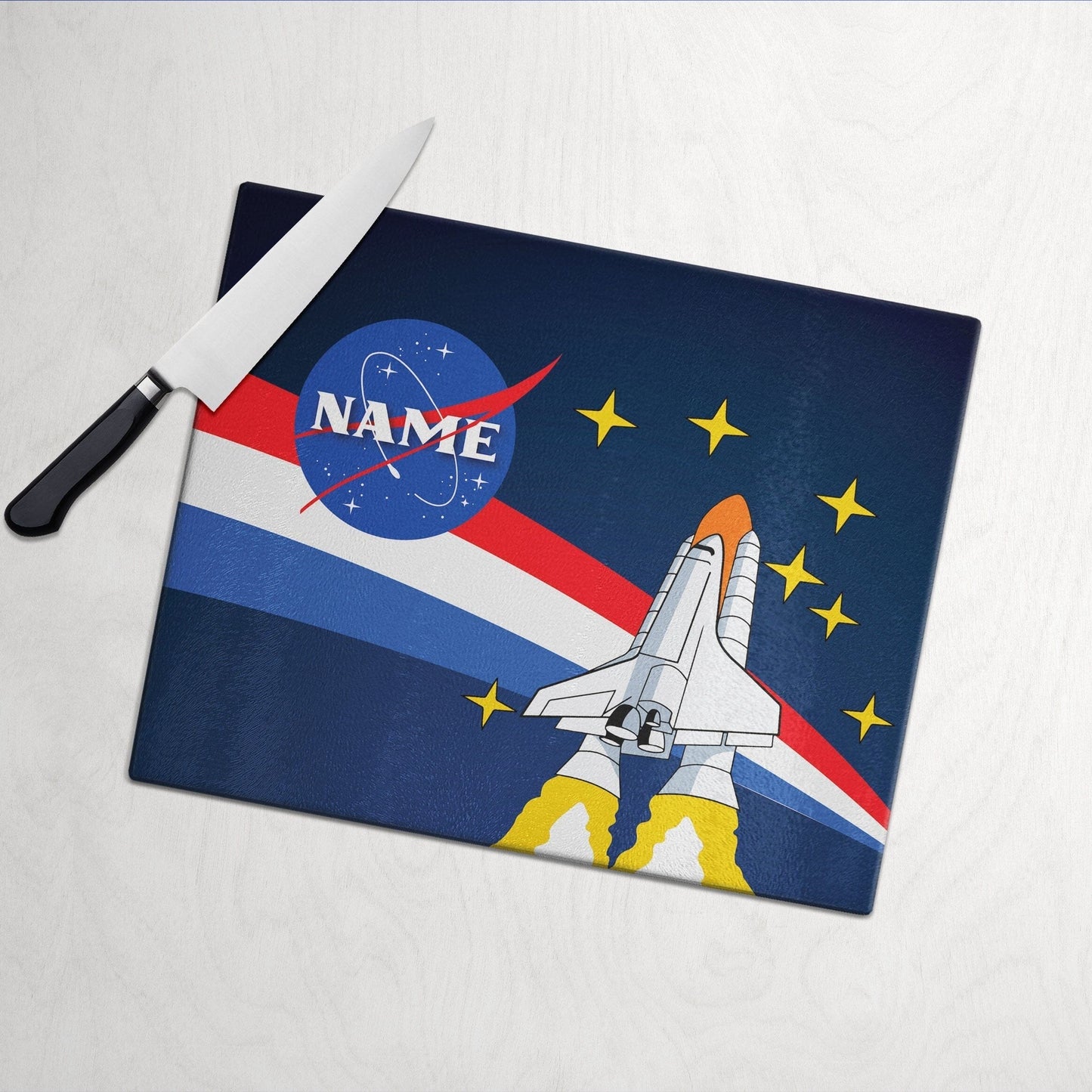 A glass cutting board with nasa launch design on a white wooden surface and knife on side